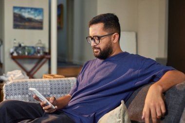 Evdeki koltukta otururken gözlük takan İspanyol bir adam. Gözlüklü bir adam cep telefonuna bakarken koltukta dinleniyor. Kanepede oturan ve akıllı telefondan fotokopi çeken karışık ırkçı adam..