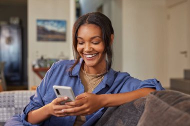 Evdeki kanepede otururken akıllı telefon kullanan gülümseyen Hintli kadın. Mutlu melez kadın rahat bir ortamda cep telefonuyla rahatlatıcı bir anın tadını çıkarıyor. Neşeli kız günlük kıyafetleriyle internette cep telefonuyla sörf yapıyor.. 