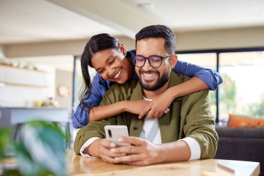 Akıllı telefona bakarken gülümseyen mutlu Hintli çift. Güzel, genç bir kadın evde akıllı telefon kullanırken kocasına arkadan sarılıyor. Orta Doğulu çift rahat ve modern bir evde kucaklaşırken yemeklerini internetten sipariş ediyorlar.