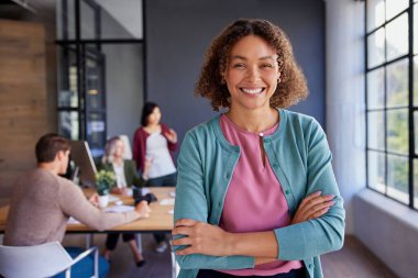 Modern bir ofiste arka planda ekibiyle gülümseyen kendine güvenen bir iş kadını. Kollarını kavuşturup gururla duran, iş yerinde liderlik ve pozitifliği somutlaştıran profesyonel bir Afrikalı kadın. İşbirliği yaparak genç bir bayanı güçlendirdi.