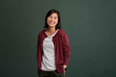 Kendine güveni olan, güler yüzlü, günlük kıyafetleriyle yeşil duvarda izole edilmiş, fotokopi alanı olan bir kız. Dost canlısı Çinli genç bayan elleri ceplerinde sıradan bir poz vererek gülümsüyor. Rahat giyinmiş mutlu bir kadın, açgözlülüğe karşı gülümsüyor.