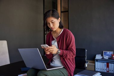 Modern bir ofiste dizüstü bilgisayarla çalışırken telefonuna bakan dalgın Çinli genç bir kadın. Endişeli Asyalı iş kadını telefonda çalışırken masada oturuyor. Bir e-posta gönderirken dijital aygıtlarla birden fazla iş yapan düşünceli iş kızı. 