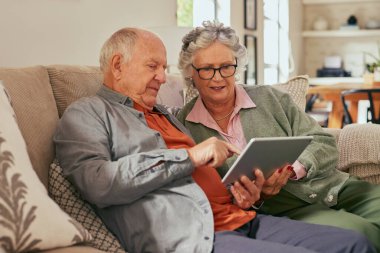 Evde dijital tablet kullanırken kanepede oturan yaşlı çift. Yaşlı bir adam ve yaşlı bir kadın bir tablette içeriği keşfediyorlar. Rahat bir emekli kadın ve yaşlı kocası bir dokunmatik ekran aygıtı, bağlantı ve iletişim kullanarak bir anı paylaşıyorlar.