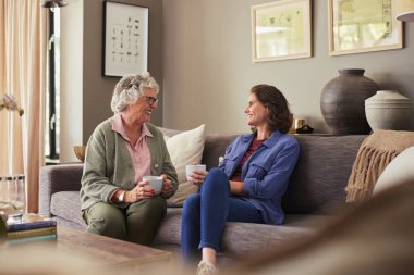 Kıdemli gülümseyen kadın ve yetişkin kız evde sıcak bir kahve fincanı içerken sohbet ediyorlar. Yaşlı anne ve genç kadın kanepede dinlenirken güzel bir sohbet ediyorlar. Mutlu yaşlı kadın ve kız kanepede oturup sohbet ediyorlar..
