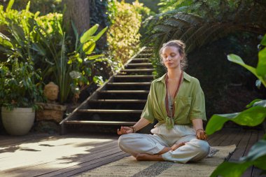Huzurlu bir kadın yemyeşil yemyeşil bir ortamda meditasyon pozunda oturuyor, sağIık merkezinde, farkındalığı ve iç huzuru simgeliyor. Açık havada meditasyon yapıyorum. Sakin ve merkezci genç bir kadın sabah zihni boyunca doğanın iyileştirici enerjisini kucaklıyor.