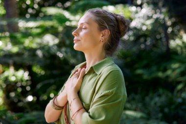 Gözü ve kolları göğsünde kapalı bir kadın yaz kampının tadını çıkarırken farkındalık egzersizi yapıyor. Kadınlar barışçıl bir inziva ortamında meditasyon ve duygusal temeller üzerinde çalışırlar. Sakin bir kız ellerini nazikçe kalbinin üzerinde tutar, memnuniyeti kucaklar.