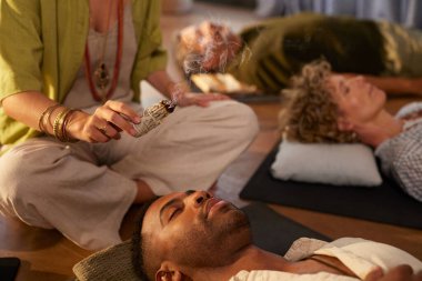 Saglik inzivasindaki enerjik oturum sirasinda bir kadin adaçayı ile kirletme ayini yapiyor. Sakin ve kutsal mekanda ruhani temizlik uygulaması. Enerji ve aura 'yı temizlemek için leke sürmek, beyaz adaçayı yakmak. Bir grup farklı insan arka planda yatıyor.