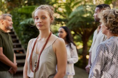 SağIık inzivası sırasında arkasında bir grupla güvenle dışarıda duran genç bir kadın. Etrafı yeşil ve çok ırklı insanlarla çevrili kameraya bakan barışçıl sarışın bir kızın portresi. Doğada sakin bir anın tadını çıkaran kadın sağlık merkezidir.