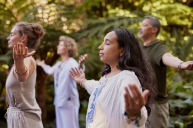 Dikkatlice meditasyon yapan bir grup insan gözleri kapalı ve kolları açık. Barışçıl bir açık hava ortamında duran bireyler, nefes ve iç denge üzerine odaklanıyorlar. Meditasyonla doğayla bağlantı kuran katılımcılar sağIık geri çekiliyor.