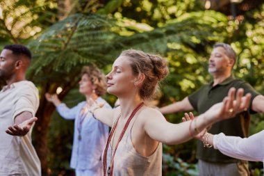 Huzurlu bir orman ortamında kollarını açarak dikkatli nefes alma alıştırması yapan çeşitli bir grup. Mulietnik insanlar açık havada, gözleri kapalı meditasyon yapıyor, iyileşme ve bağlantı kurmaya odaklanıyor. Doğallıkla çevrili bir sağlık inzivasına katılan katılımcılar