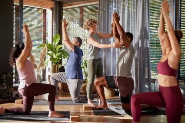 Yoga eğitmeni, bir grup çok ırklı yetişkinin ders sırasında egzersiz yapmalarına yardım ediyor. Yoga pratiği yapan insanlar, kadın bir öğretmenin rehberliğiyle içeride poz veriyorlar. Çeşitli gruplar yoga öğreniyor. Barışçıl ve parlak bir stüdyoda, savaşçı pozunda..