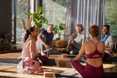 Bir grup melez yetişkin güneşli bir stüdyoda yoga yaptıktan sonra rahatlayıp sohbet ediyorlar. Gülümseyen çeşitli insanlar yoga dersinden sonra sohbet etmekten hoşlanırlar. Fitnes 'den sonra bir grup mutlu erkek ve gülümseyen kadın spor salonunda sohbet ediyorlar.