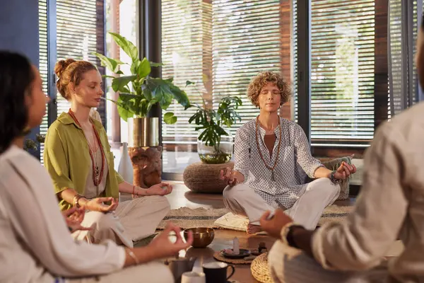 Bir grup insan, kapalı bir sağlık merkezinde birlikte meditasyon yapıyor. Sakin, olgun bir kadın bacak bacak üstüne atmış, gözleri kapalı, farkındalık ve iyileşme pratiği yapıyor. Yoga dersi sırasında çeşitli insanlar oturup meditasyon yapıyor..