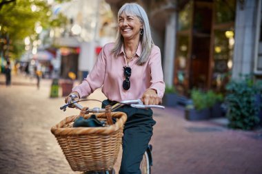 Gülümseyen yaşlı kadın büyük bir gülümsemeyle bir şehir caddesinde bisiklet sürüyor. Fotokopi alanı olan bir şehirde bisikletle gezen mutlu olgun bir kadın. Neşeli bayan Avrupa 'da huzurlu bir bisiklet yolculuğunun tadını çıkarıyor..