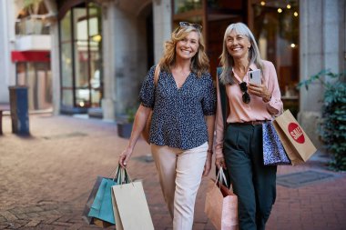 Gündelik kıyafet giymiş iki yetişkin kadın satış promosyonu sırasında alışveriş çantalarıyla mutlu bir şekilde yürürler. Üst düzey yaşlı arkadaşlar birlikte dışarıda alışveriş yapmanın tadını çıkarırlar. Alışveriş torbaları ve fotokopi yeriyle sokakta yürüyen mutlu, gülen kadınlar..