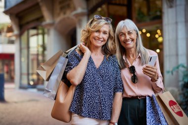 Birlikte alışveriş yaptıktan sonra gülümseyen iki olgun kadın. Gülümseyen son sınıf hanımlarının portresi ellerinde alışveriş torbalarıyla mutlu poz veriyor. Yaşlı kadınlar kameraya bakarken neşeli bir alışveriş gününün tadını çıkarırlar..