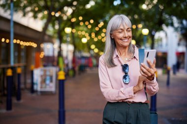 Yaşlı bir kadın, şehir caddesinde yürürken telefonuna bakarken gülümser. Akıllı telefonlu ve fotokopi çekilen bir görüntüde olgun, güzel bir kadın. Güzel aydınlatılmış bir sokakta telefon mesajlarını kontrol eden yaşlı bir kadın..