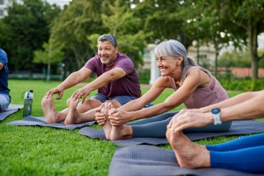 Son sınıflar şehir parkındaki yoga minderleri üzerinde birlikte esneme egzersizleri yapıyorlar. Yaşlı insanlar ayak parmaklarına dokunmak için öne eğilirken gülümserler. Yoga sırasında parkta spor yapmak için bir grup kadın ve erkek..