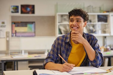 Erkek öğrenci elinde kalem tutarken gülümsüyor ve sınıfta fotokopi çekerek düşünüyor. Sıradan bir genç öğrenci masada oturuyor, çalışıyor ve sınıfta kendinden emin hissediyor. Lisede gururlu ve memnun bir genç ders sırasında gözlerini kaçırıyor.
