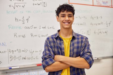 Beyaz tahtanın önünde dikilirken güvenle gülümseyen çok ırklı bir öğrenci. Genç öğrenci sınıfta çapraz kollarla denklemleri çözdükten sonra gururla poz verir. Latin mutlu adam, matematik ya da geometri problemini çözdükten sonra gururla kameraya bakıyor.