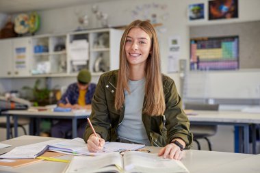 Kitaplar ve notlarla çalışırken gülümseyen mutlu bir üniversite öğrencisi. Öğrenci not defterine yazar ve kameraya bakar. Ortaokulda gülümseyen sıradan bir genç kadının portresi.. 