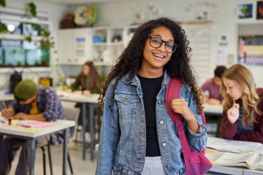 Sırt çantalı gülümseyen Afrikalı Amerikalı öğrenci derse başlamaya hazır. Mutlu gülümseyen genç kız lisede arka planda ders çalışan sınıf arkadaşlarıyla güven içinde poz veriyor. Parlak ve neşeli öğrenci geri dönmenin tadını çıkarıyor.