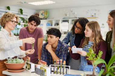 Sınıf etkinliği sırasında bir grup etnik çeşitliliğe sahip öğrenciye deney tüpünde numune gösteren olgun bir fen öğretmeni. Liseli çocuklar ve kızlar bitki örneğiyle ilgili biyoloji deneyini gözlemlemek için masanın etrafında toplandılar. Bir grup genç üniversite sınıf arkadaşı.