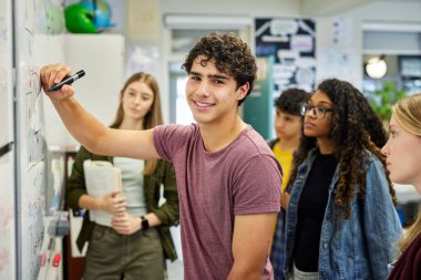 Sınıf arkadaşlarına bir konsepti açıklarken beyaz tahtaya yazı yazan zeki bir öğrenci. Lisede matematik dersi sırasında beyaz tahtaya aritmetik denklemin mutlu çocuk genç yazım çözümü. Sınıf arkadaşlarının çözümü anlamasına yardım eden genç adam.