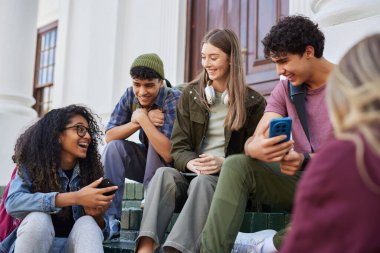 Kampüs basamaklarında oturan, gülümseyen ve akıllı telefonlarındaki içeriği paylaşan çok ırklı bir grup öğrenci. Mutlu erkekler ve gülümseyen kızlar cep telefonuyla video izleyip eğleniyorlar. Bir grup genç arkadaş birlikte otururken telefon uygulamasını kullanarak komik şeyleri paylaşıyorlar. 