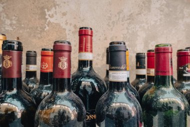 Old bottles of red and white wine full of dust with a wall with the paintwork ruined by time in the background. Italy