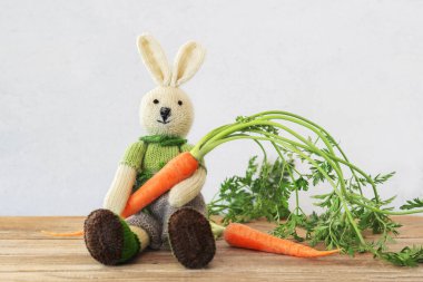 Knitted cute bunny sitting on wooden table holding a fresh carrot with copy space