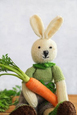 Knitted cute bunny sitting on wooden table holding a fresh carrot with copy space