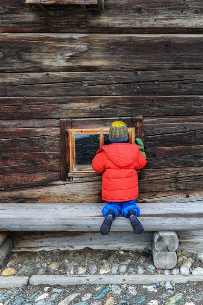 一个穿着红色夹克的小孩跪在地上 好奇地凝视着一个木制谷仓的窗户 试图探索他所处的环境 — 图库照片