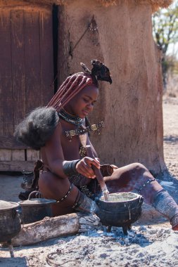 Damaraland, Namibya - 14 Temmuz 2024: Genç bir Himba kadını kulübesinin önünde mısır lapası pişiriyor. Himba halkı Kuzey Namibya 'da yaşayan yerli bir kabiledir.