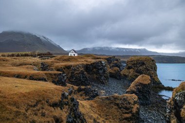 Arnarstapi kıyıları. Beyaz bir evi olan tuhaf kaya oluşumuyla Snaefellsnes dağı arka planda duruyor.