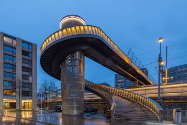 The pedestrian bridge with illumination over the rail way at the Zurich main station at the blue hour. A train is passing by under. clipart