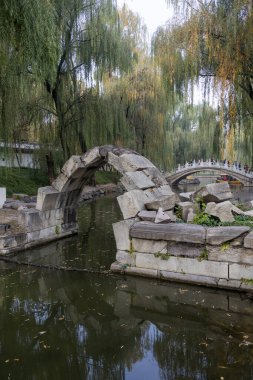 Beijing, China- 3 Nov 2024: Ruin stone bridge inside Yuanmingyuan Park in Beijing, China. It is the only one stone bridge left in Yuanmingyuan, a small arch over a narrowing in the stream. clipart