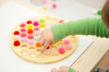 3 yaşındaki bir çocuk bir Rus hamur makinesiyle oynuyor ve ponponları deliklere sokuyor. Plastik bir torbada renkli iplik topları. Montessori konsepti. Erken eğitim, renk oyunu, iyi motor beceriler..