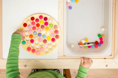 3 yaşındaki bir çocuk bir Rus hamur makinesiyle oynuyor ve ponponları deliklere sokuyor. Plastik bir torbada renkli iplik topları. Montessori konsepti. Erken eğitim, renk oyunu, iyi motor beceriler..