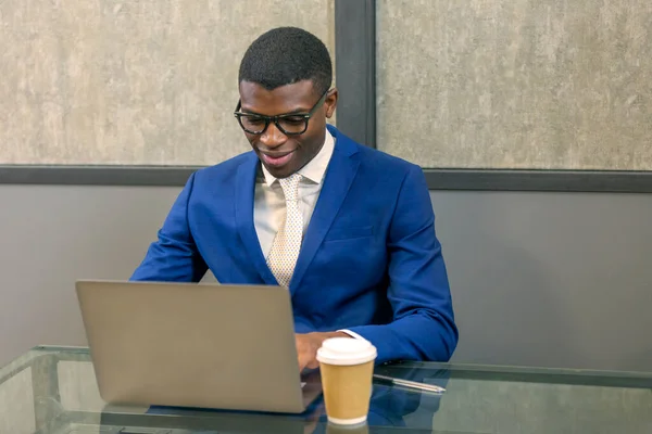 Smiling African business man wearing suit, eyeglasses  working on laptop from  office. Black man   using computer, sitting at desk watching webinar
