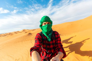 happy nomad man dress of red and green taking a selfie during camel riding in Sahara Desert, Merzouga, Morocco