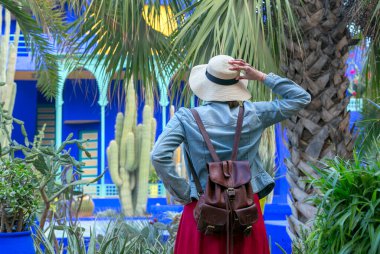 Şapkalı ve sırt çantalı genç bir kadın bambu bir patikada elinde şapkasıyla renkli tencerelerle dikiliyor. Jardin Majorelle. Marakeş, Fas