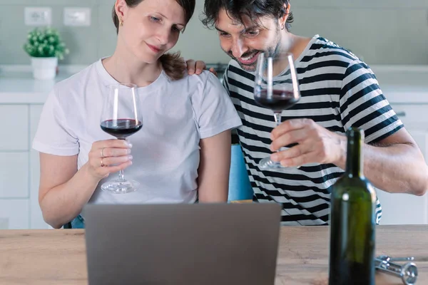 Virtual party concept. Smiling  couple sitting at the table and having online video call with friends or family using laptop, drinking wine and toasting, celebrating together at home