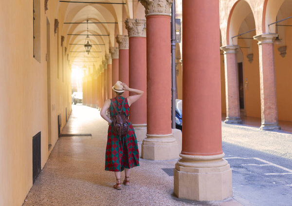 Young female tourist dressed casually standing with hat in the famous arched galleries in Bologna city in Italy. Bologna is student city and home to the oldest university in the world