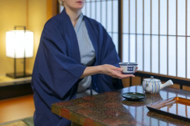 Japon elbiseli genç beyaz kadın ryokan odasındaki masada oturmuş çay içiyor.