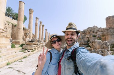a young couple with hat taking a selfie while enjoying the beautiful landscape of the ancient ruins of Jerash in a sunny day