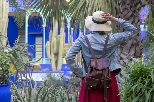 stock image A young woman in a  hat with and  backpack is standing on a bamboo alley track with colorful pots and holding her hat in her hands. Jardin Majorelle. Marrakech, Morocco