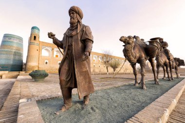 Khiva, Özbekistan - Mart - 29 - 2024: İpek Yolu boyunca çeşitli mallar taşıyan antik bir karavanın heykel grubu ve eski kent duvarları önünde geleneksel Özbek elbiseli iki adam