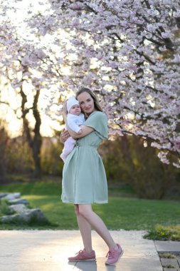 Güzel genç bir anne bebeğiyle birlikte bir bahar parkında yürüyor. Annesinin kollarında küçük bir çocuk. Yenidoğan annesiyle oynuyor.