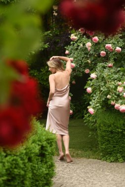 Pembe elbiseli, uzun sarı saçlı güzel bir genç kadın. Güllerin açtığı bahçede bir kadın